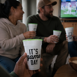 A person holds a white foam cup with green rim and black text reading 'IT'S game DAY' in the foreground, while two other people sit in a living room holding matching cups. A TV is visible in the background.