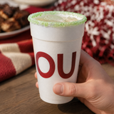 A hand holding a white foam cup with a green sugar-rimmed edge and large red letters spelling 'OU' on the front, set on a wooden table with a blurred festive background.