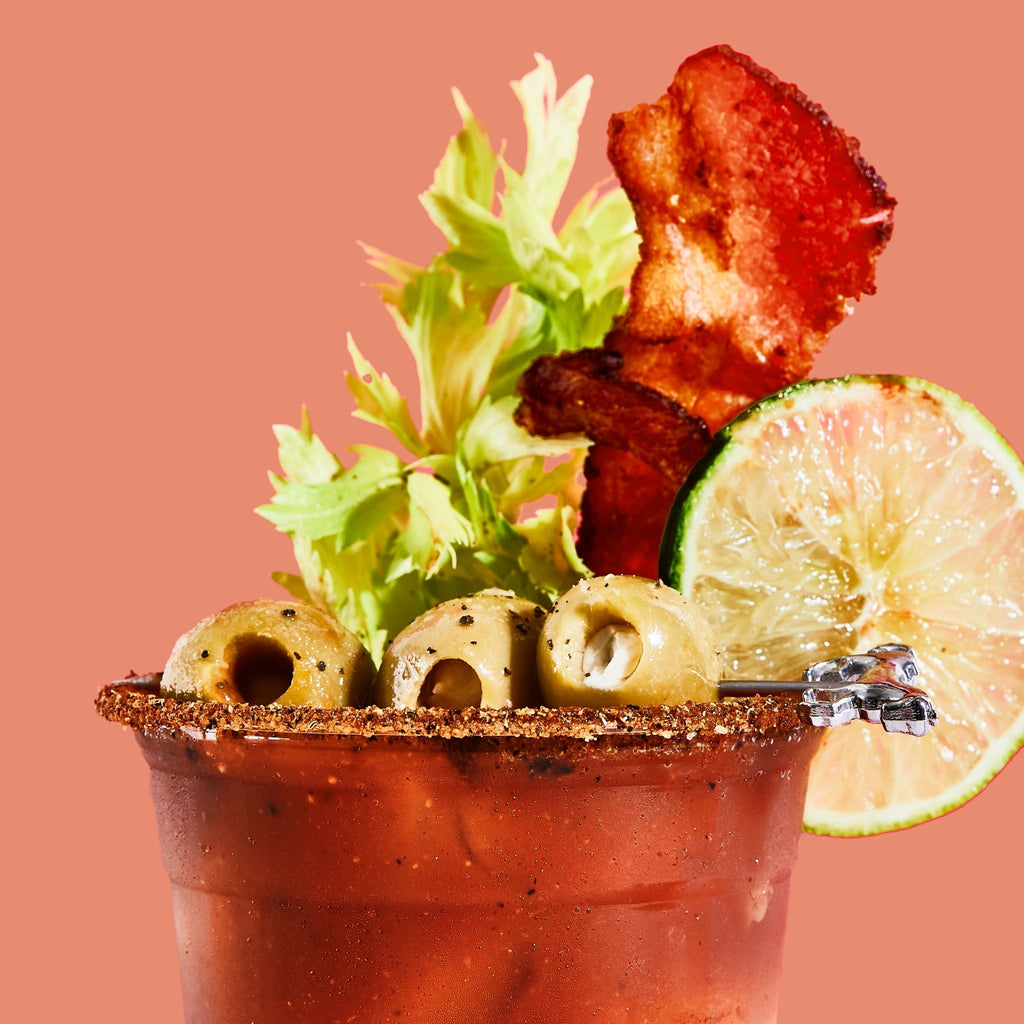 Bloody mary cocktail with bacon, lime, and olives on a pink background