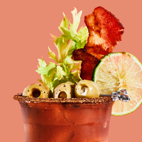 Bloody mary cocktail with bacon, lime, and olives on a pink background