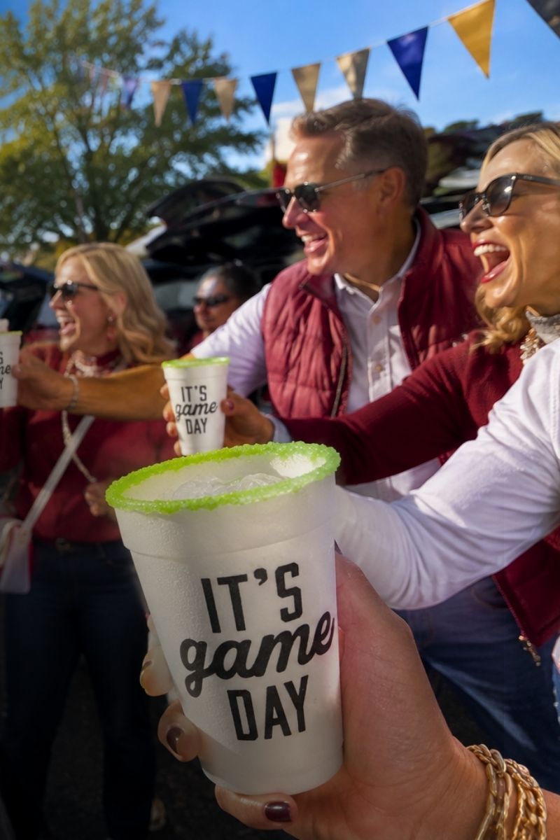 A person holds a white paper cup with 'IT'S game DAY' printed in black script and a bright green Salt & Lime rim while smiling people at an outdoor event cheer in the background.